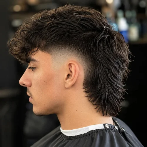 Young man in side profile showing a textured curly modern mullet with a clean skin fade around the ear and longer layered hair at the back, photographed in a barbershop.